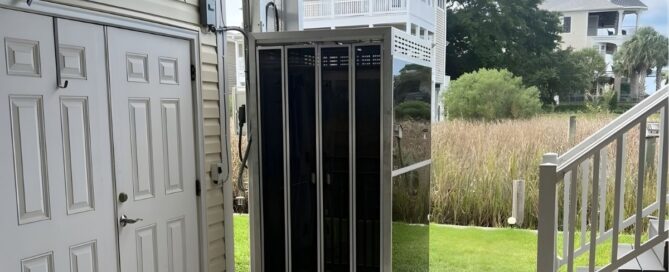 Outdoor home elevator next to a beige siding house, surrounded by grass and stairs; white multi-story houses in the background.