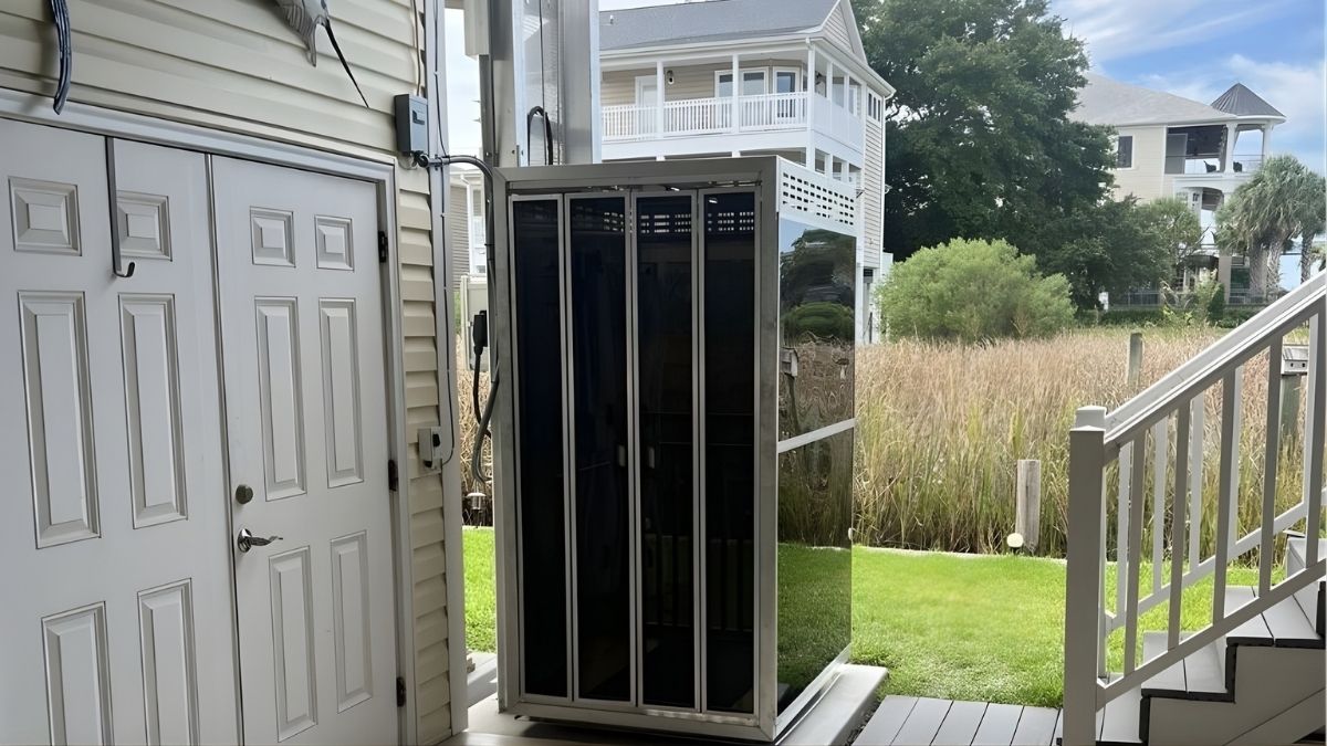 Outdoor home elevator next to a beige siding house, surrounded by grass and stairs; white multi-story houses in the background.
