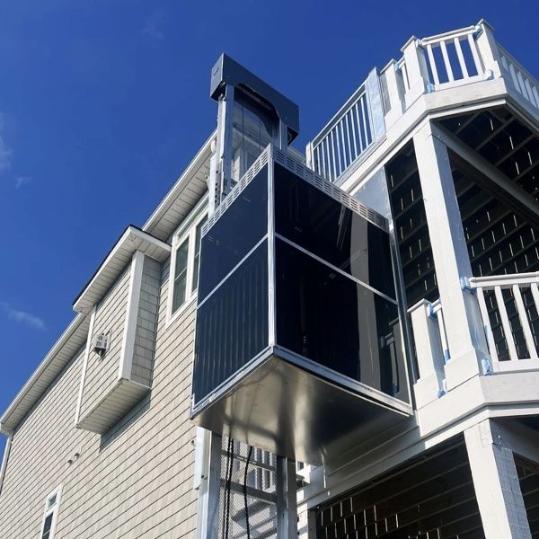 Outdoor installation of a modern home elevator on a multi-story beige house with white railings against a clear blue sky.