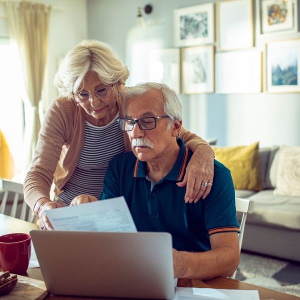 Elderly couple reviewing documents together at a dining table with a laptop, discussing home elevators. Cozy living room setting.