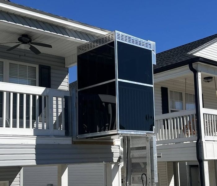Modern home elevator outside a two-story house, connecting upper balcony to ground level, under a clear blue sky.