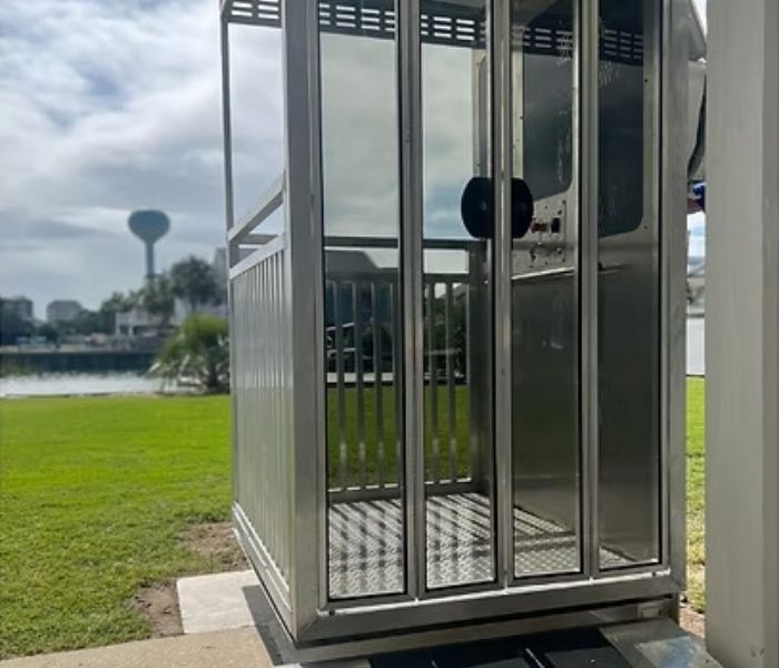 Modern glass home elevator with metal frame, situated outdoors on a sunny day, overlooking green lawn and distant water tower.