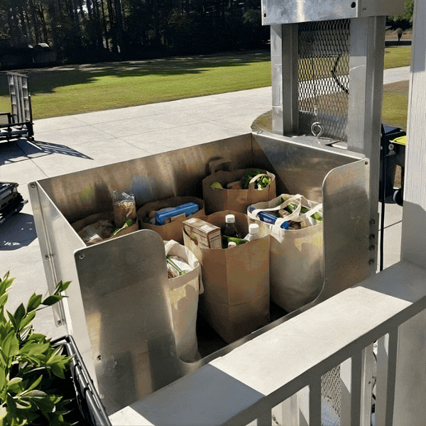 Exterior Dumbwaiter Groceries in paper bags transported by a home elevator on a sunny patio, with a grassy lawn in the background.