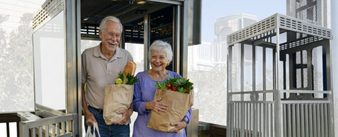 Smiling elderly couple exits a home elevator, carrying grocery bags filled with fresh produce.