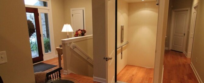 Well-lit foyer with wooden floors, open home elevator, staircase, and decorative table with lamp and flowers.