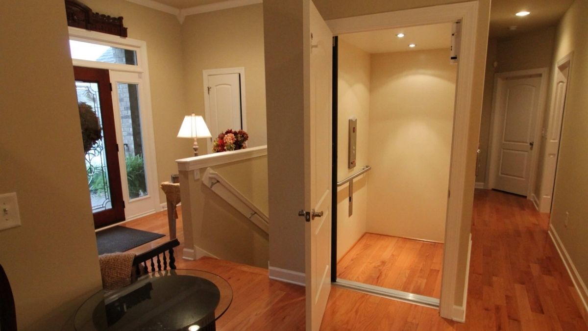 Making Your Home Work for You Long-Term Well-lit foyer with wooden floors, open home elevator, staircase, and decorative table with lamp and flowers.